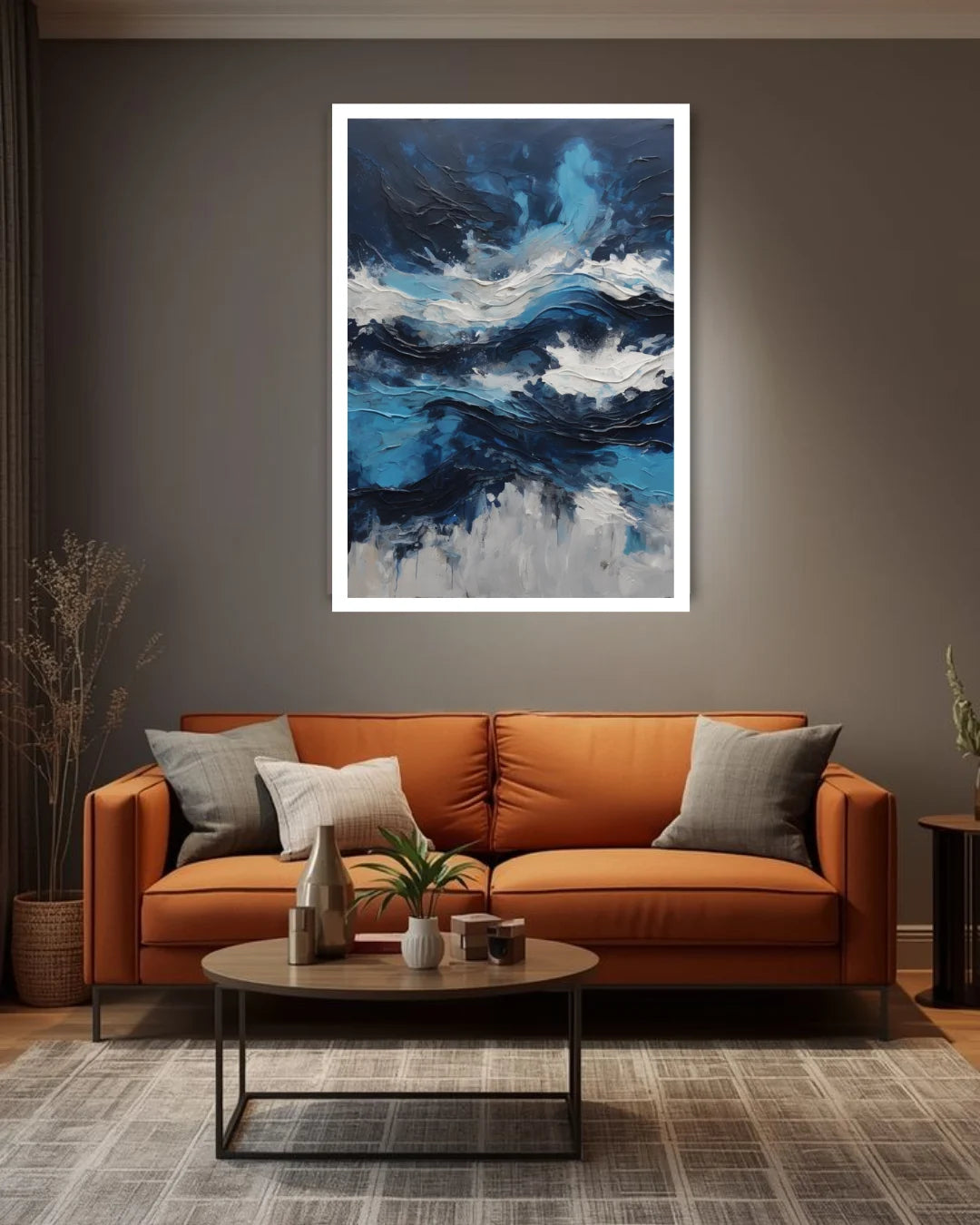 A framed abstract canvas print featuring textured blue, white, and black brushstrokes, displayed on a dark gray wall above a modern orange-brown leather sofa in a styled living room.