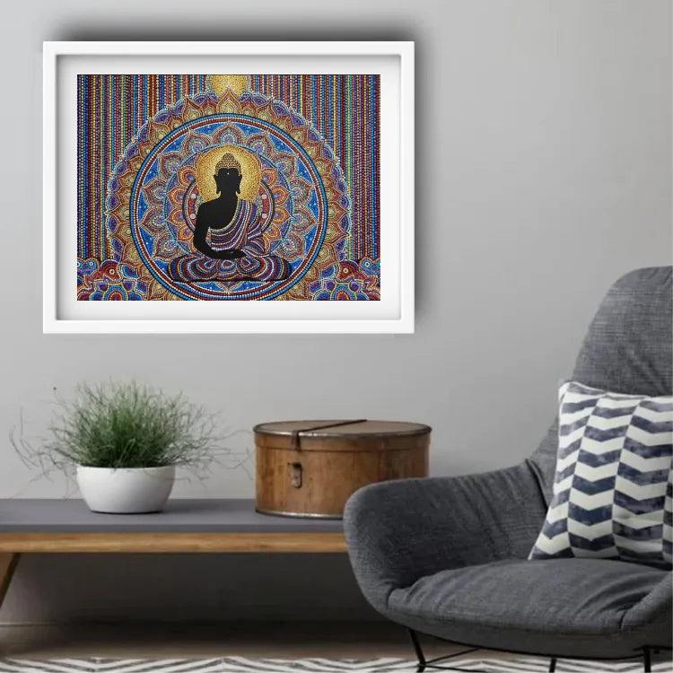 A framed wall art print featuring a black seated Buddha silhouette against a highly detailed, colorful mandala background, displayed in a white frame on a gray wall above a console table with a plant and a gray armchair.