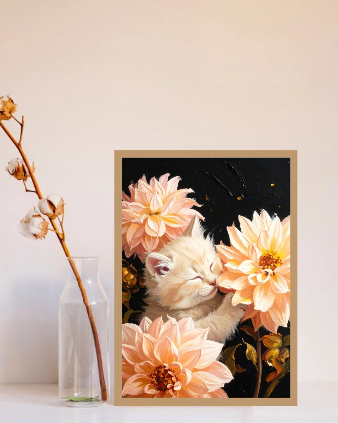 A framed art print featuring a sleeping white kitten nestled among large peach and cream dahlia flowers against a dark background, styled on a white surface next to a clear glass bottle with dried cotton stems.