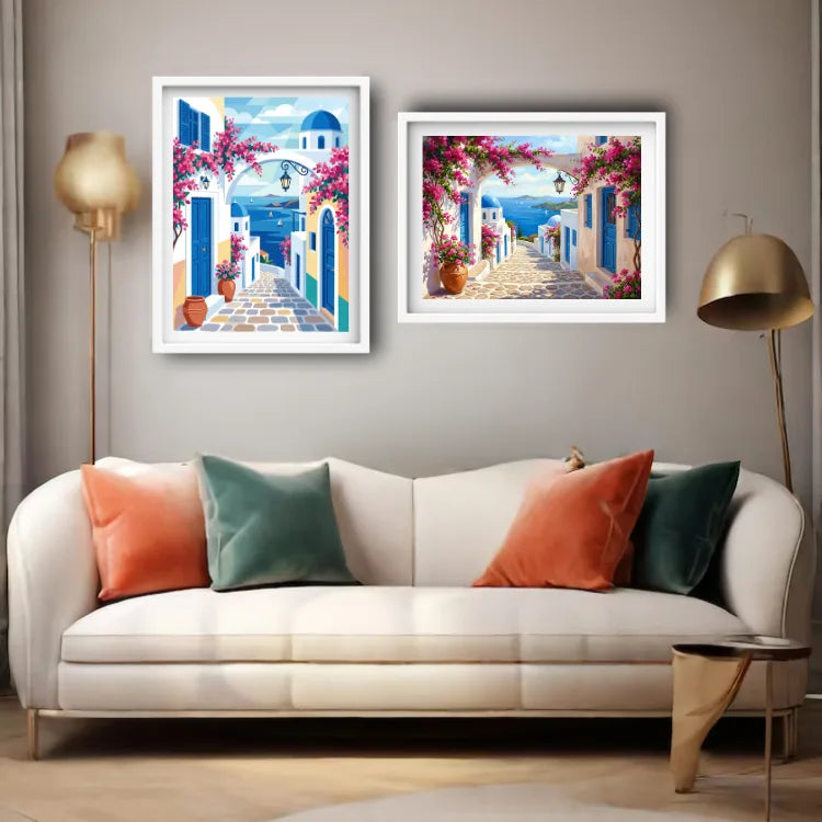 Two framed art prints depicting a Mediterranean blue-and-white coastal village scene with pink bougainvillea, displayed above a white sofa in a modern living room setting.