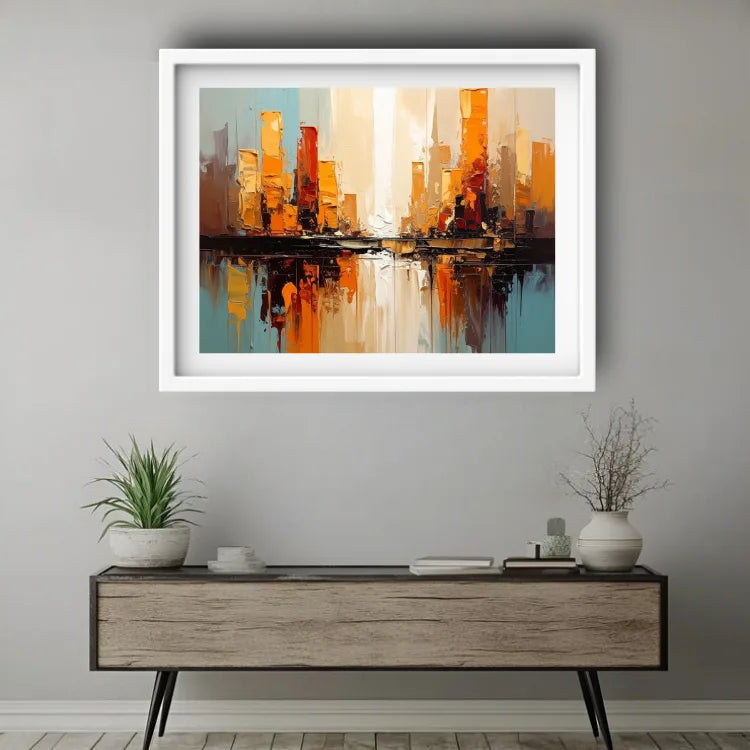 A framed abstract cityscape painting with warm orange and yellow buildings reflected in blue water, displayed on a light gray wall above a modern wooden console table with plants and decor.