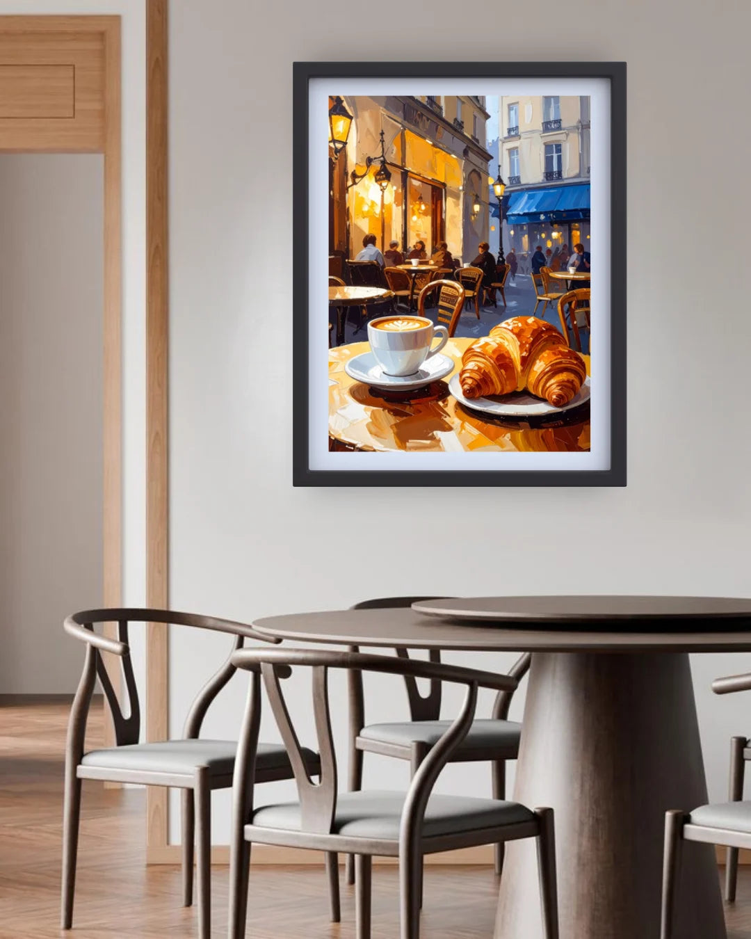 A framed wall art print depicting a Parisian cafe scene with croissants and a coffee cup, displayed above a dining table with chairs in a modern interior setting.