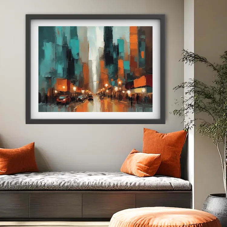 A framed abstract painting of a city street at night with teal, orange, black, and white tones, displayed on a wall above a cushioned bench with orange pillows in a styled interior room.