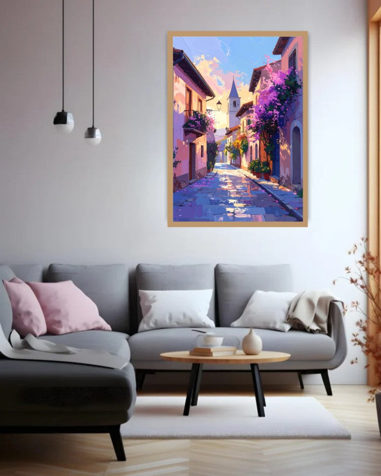 A colorful framed painting of a European village street with purple flowers and a blue sky, displayed on a light gray wall above a modern gray sofa in a living room setting.