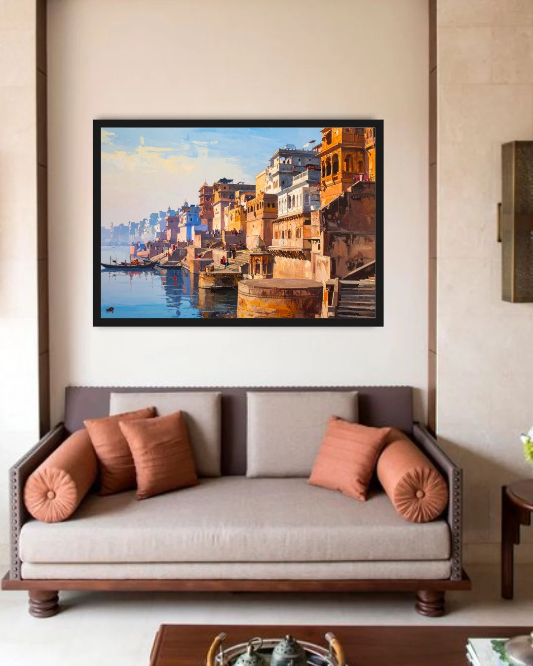 A framed painting of a colorful Indian ghats and river scene hangs on a beige wall above a modern sofa with brown and terracotta cushions in a styled living room.