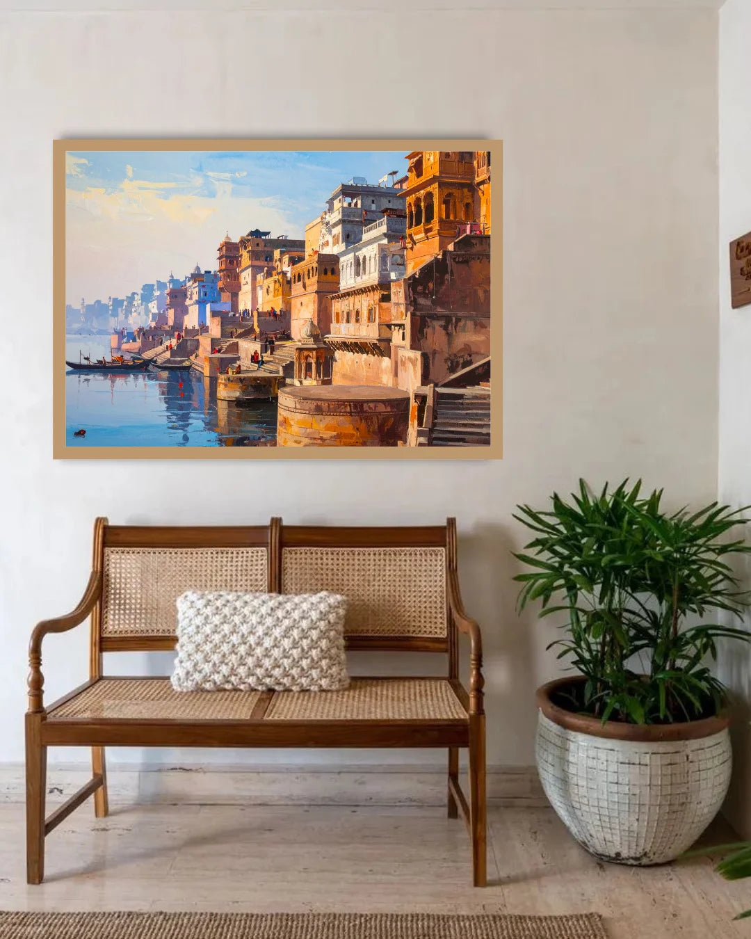 A framed painting of a ghat scene in Varan, India, hanging on a light beige wall above a wooden bench with a woven seat and a decorative pillow, next to a potted green plant.