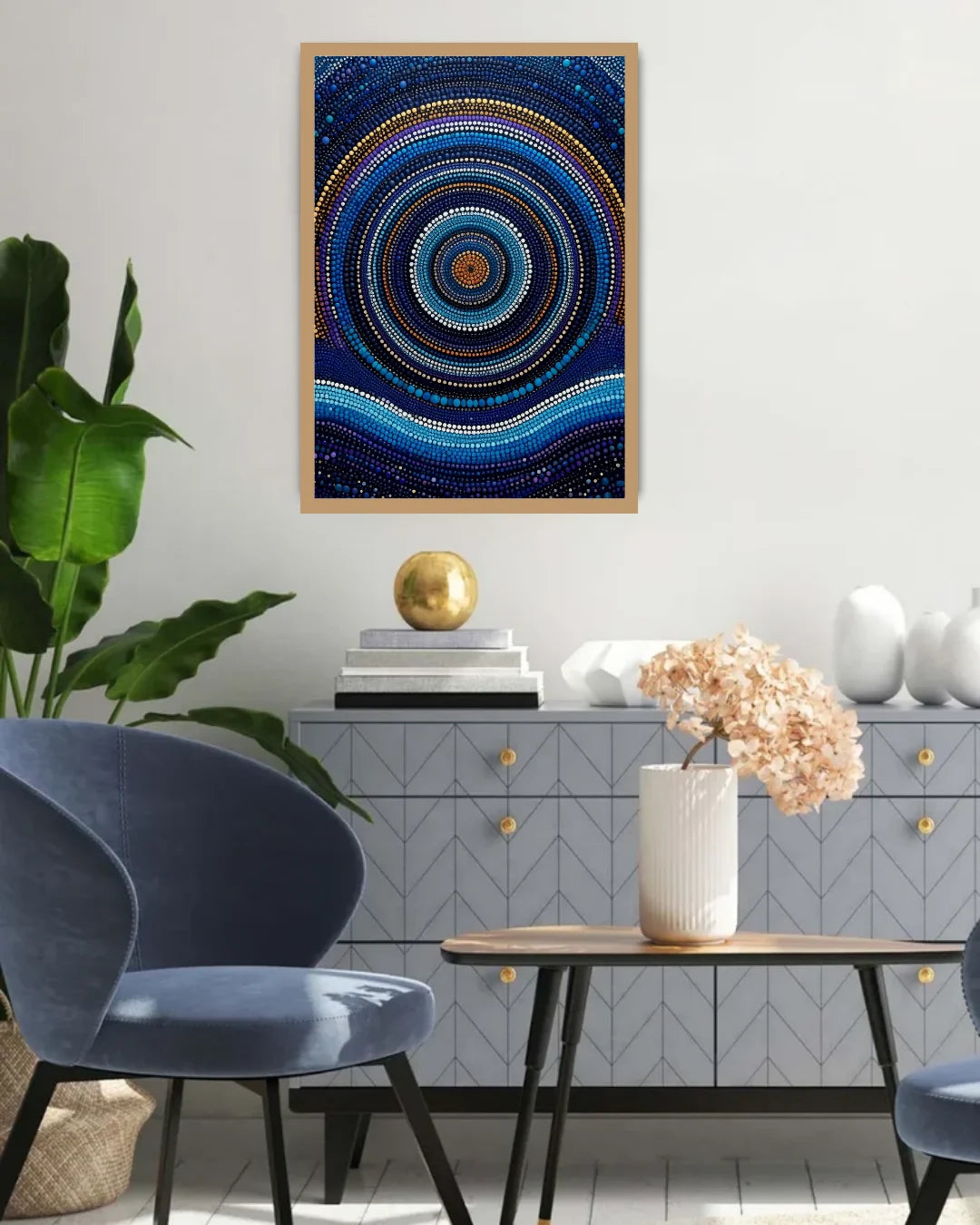 A framed abstract art print featuring concentric circles and dots in shades of blue, navy, and gold, displayed on a light gray wall in a modern living room setting.
