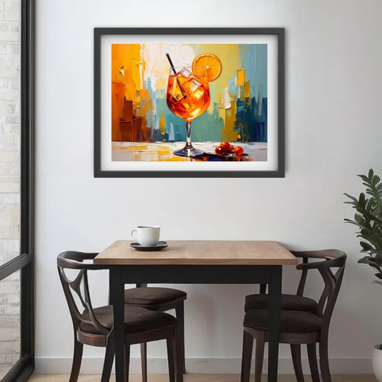 A framed abstract art print of an orange cocktail with a lemon slice, displayed on a white wall above a wooden dining table with two chairs.