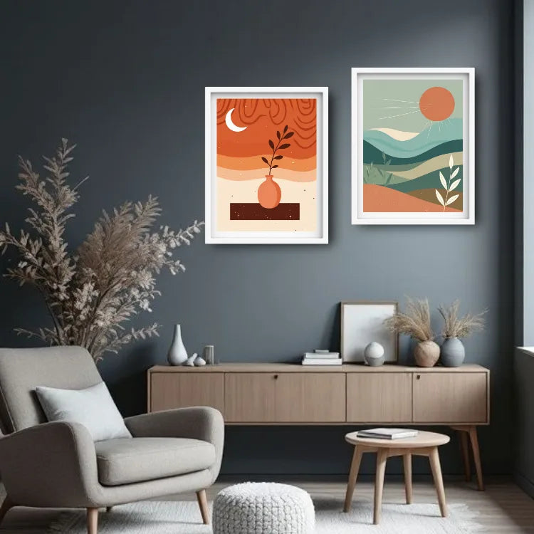 Two framed minimalist art prints hang on a dark gray wall above a light wood console table in a styled living room. The left print features orange and beige abstract waves with a small orange vase and dark branch, and the right print shows layered green and orange hills under a large orange sun.
