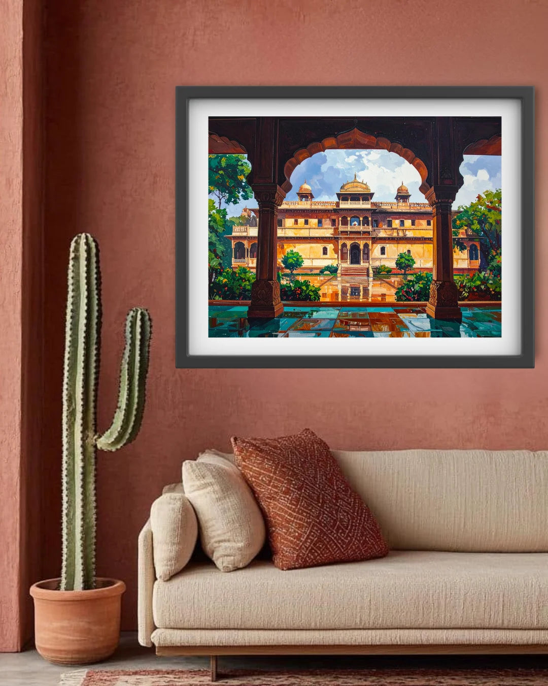 A framed art print depicting a colorful palace scene viewed through arches, displayed on a terracotta wall above a beige sofa with decorative pillows and a potted cactus nearby.