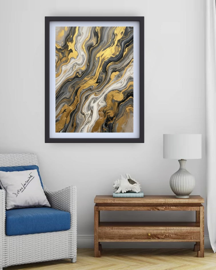 A framed abstract art print with swirling gold, black, gray, and white marble-like patterns, displayed on a light gray wall above a wooden side table and next to a white armchair with a blue cushion.