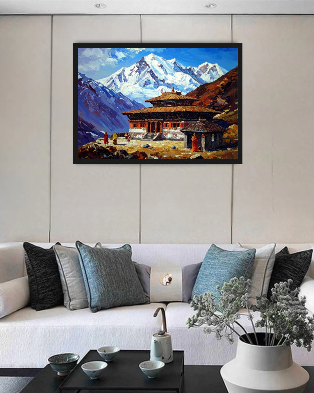 A framed landscape painting of snow-capped mountains and a traditional building, displayed on a beige paneled wall above a white sofa with decorative pillows.