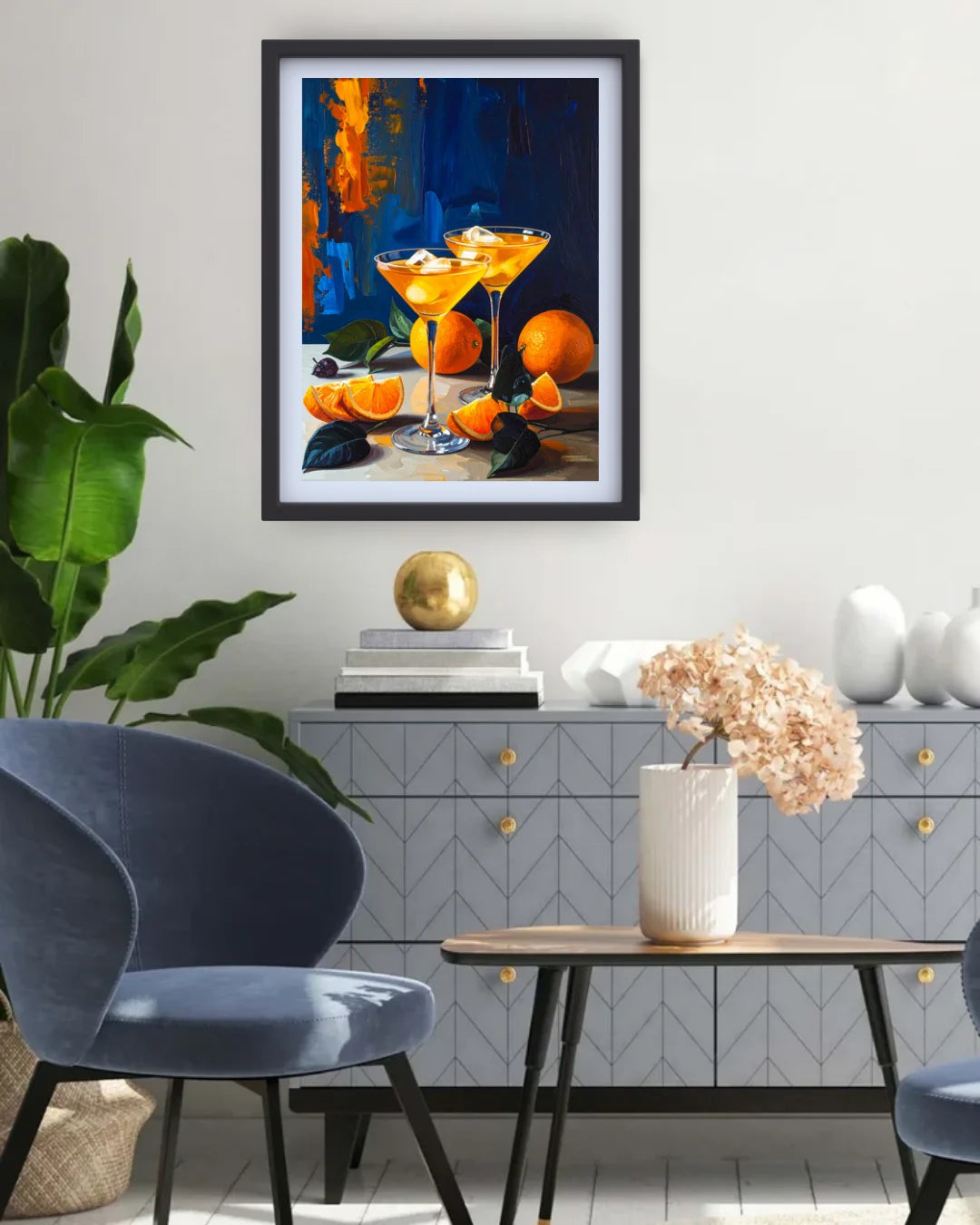 A framed art print depicting two orange Martini glasses and oranges on a table, styled in a modern living room setting above a gray sideboard.