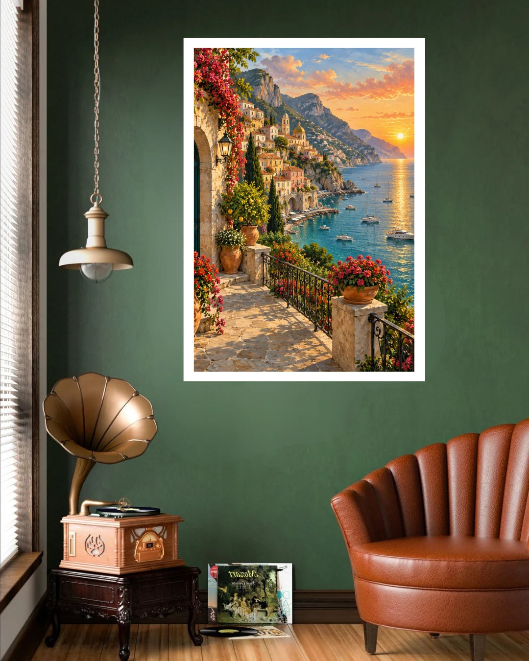 A framed vertical art print depicting a scenic Mediterranean coastal town with a sunset, hanging on a dark green wall in a styled room with a brown leather chair, a vintage gramophone, and a hanging lamp.