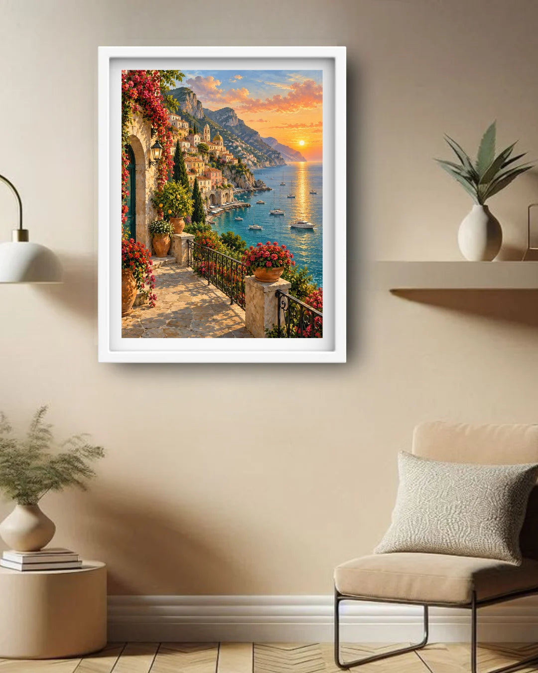 A framed art print depicting a scenic Mediterranean coastal town at sunset, hung on a beige wall in a modern living room setting with a chair, side table, and plants.
