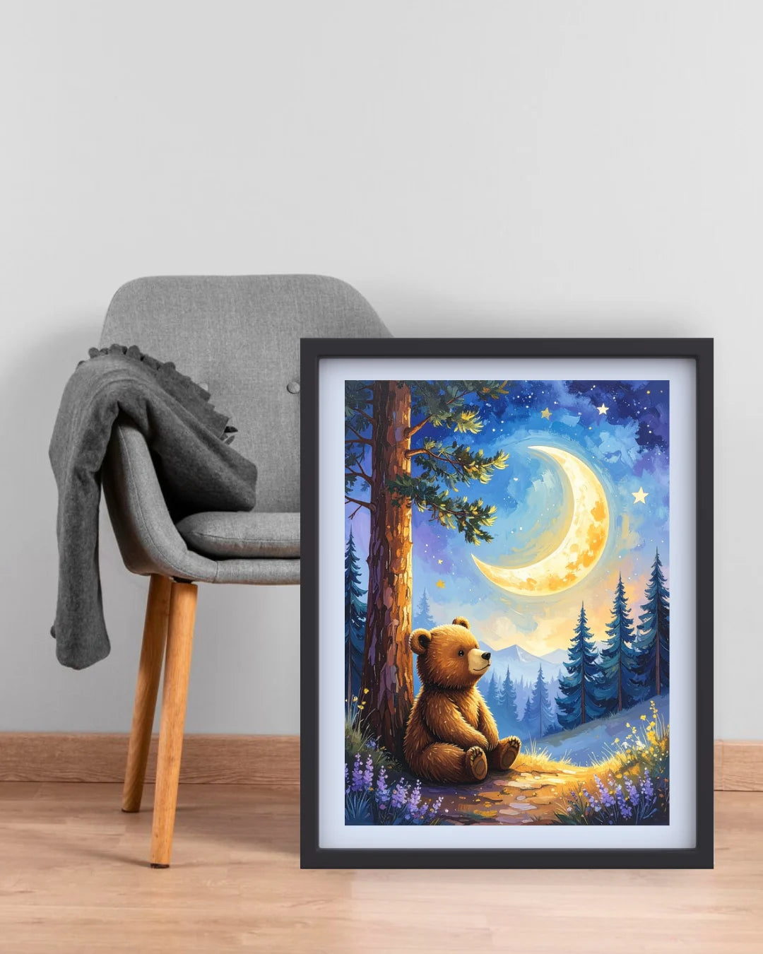 A framed art print depicting a cozy night scene with a brown bear sitting by a tree under a glowing crescent moon and stars, styled next to a gray armchair in a room setting.