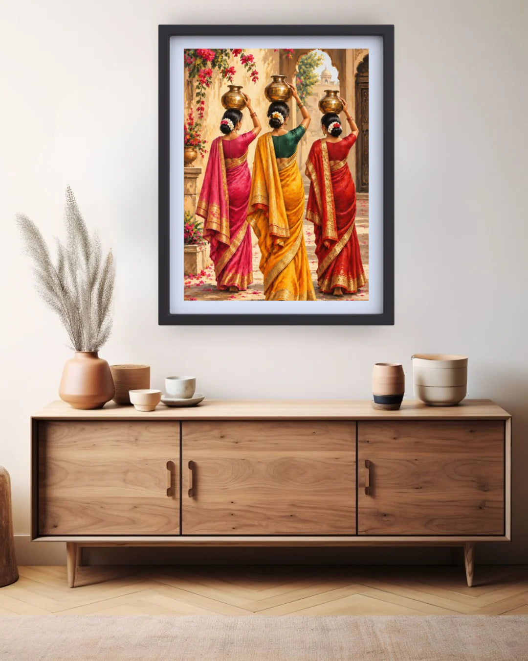 A framed art print depicting three Indian women in colorful traditional sarees carrying brass pots, displayed on a light beige wall above a wooden sideboard with decorative vases and cups.