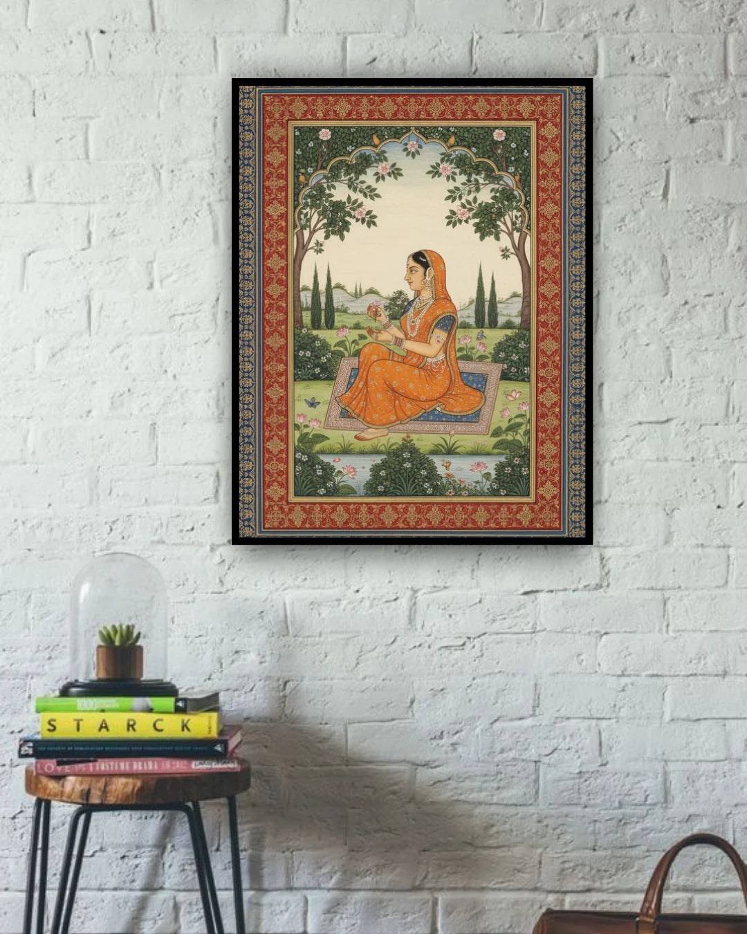 Framed artwork of a woman in an orange saree on a white brick wall with a small table and books below.