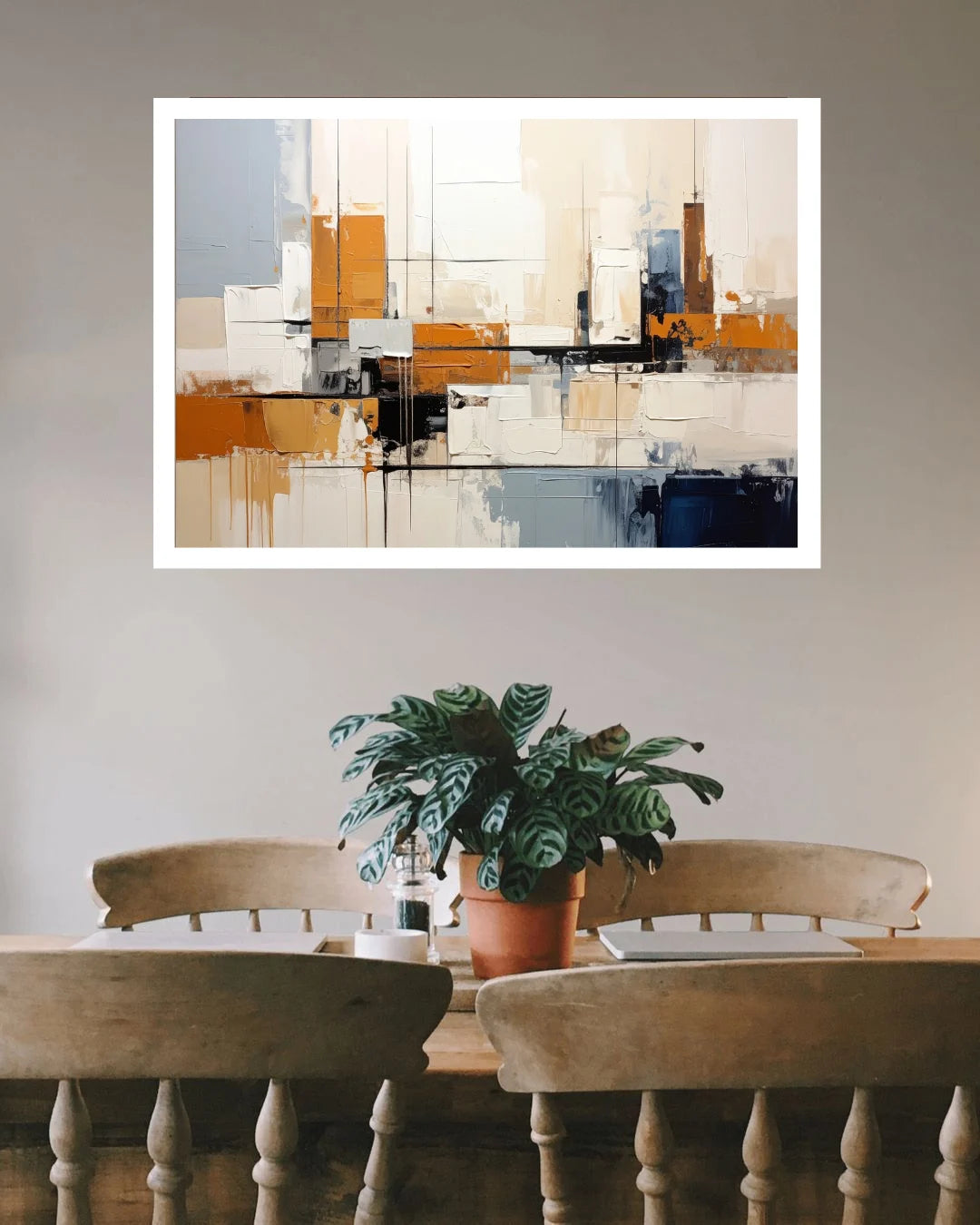 An abstract canvas wall art print with geometric blocks of white, beige, orange, gray, and blue, displayed in a white frame on a light gray wall above a dining table with chairs and a potted plant.