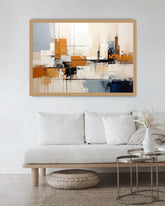 A framed abstract canvas print with geometric shapes in orange, beige, white, gray, and blue, displayed on a white wall above a modern white sofa in a bright living room.