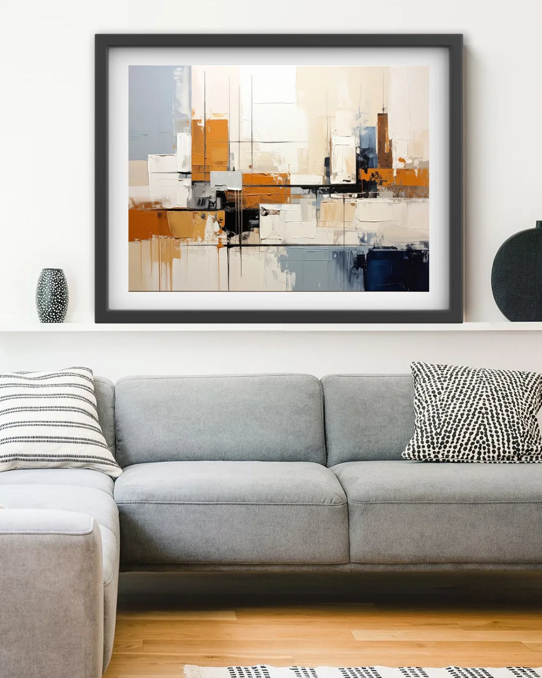 A framed abstract canvas print with a black frame and white mat, featuring geometric blocks of orange, beige, gray, and blue, displayed on a white wall above a modern gray sectional sofa.