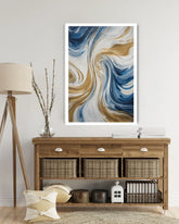 A framed abstract wall art print with swirling blue, white, and tan brushstrokes, displayed above a wooden console table in a styled room setting.
