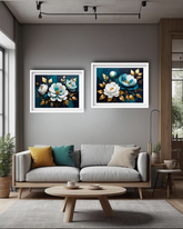 A modern living room setting featuring two square framed floral paintings with white and blue flowers on a dark teal background, hung symmetrically above a light gray sofa.