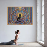 A colorful framed wall art print featuring a seated Buddha silhouette with intricate mandala-style patterns, displayed on a white wall in a bright room. A woman in athletic wear is lying on a yoga mat below, in a yoga pose.