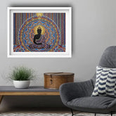 A framed wall art print featuring a black seated Buddha silhouette against a highly detailed, colorful mandala background, displayed in a white frame on a gray wall above a console table with a plant and a gray armchair.