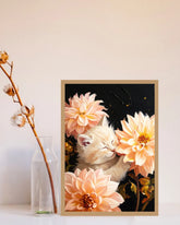 A framed art print featuring a sleeping white kitten nestled among large peach and cream dahlia flowers against a dark background, styled on a white surface next to a clear glass bottle with dried cotton stems.