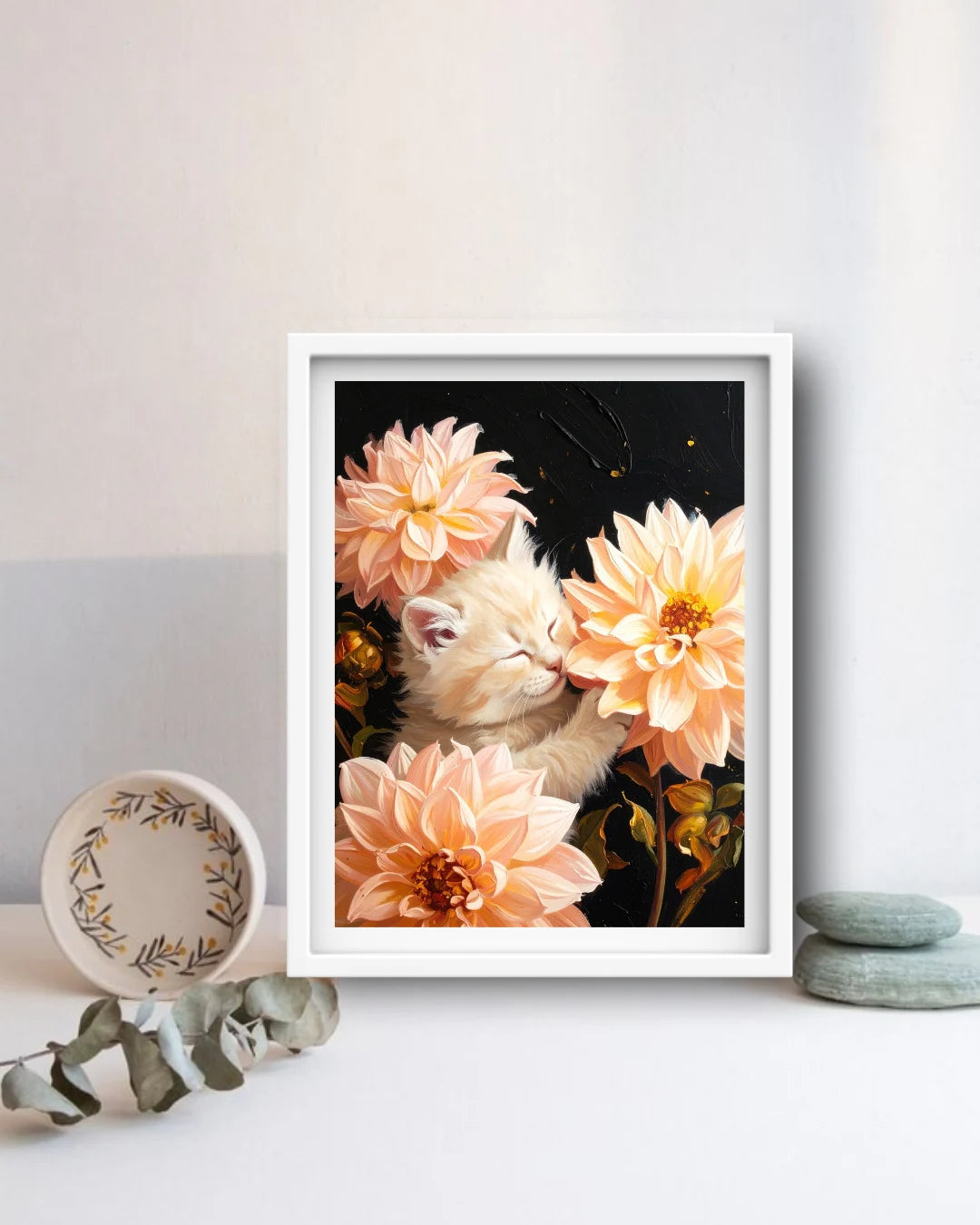 A framed art print featuring a white kitten nestled among peach and orange dahlia flowers against a dark background, styled on a white surface with decorative props.