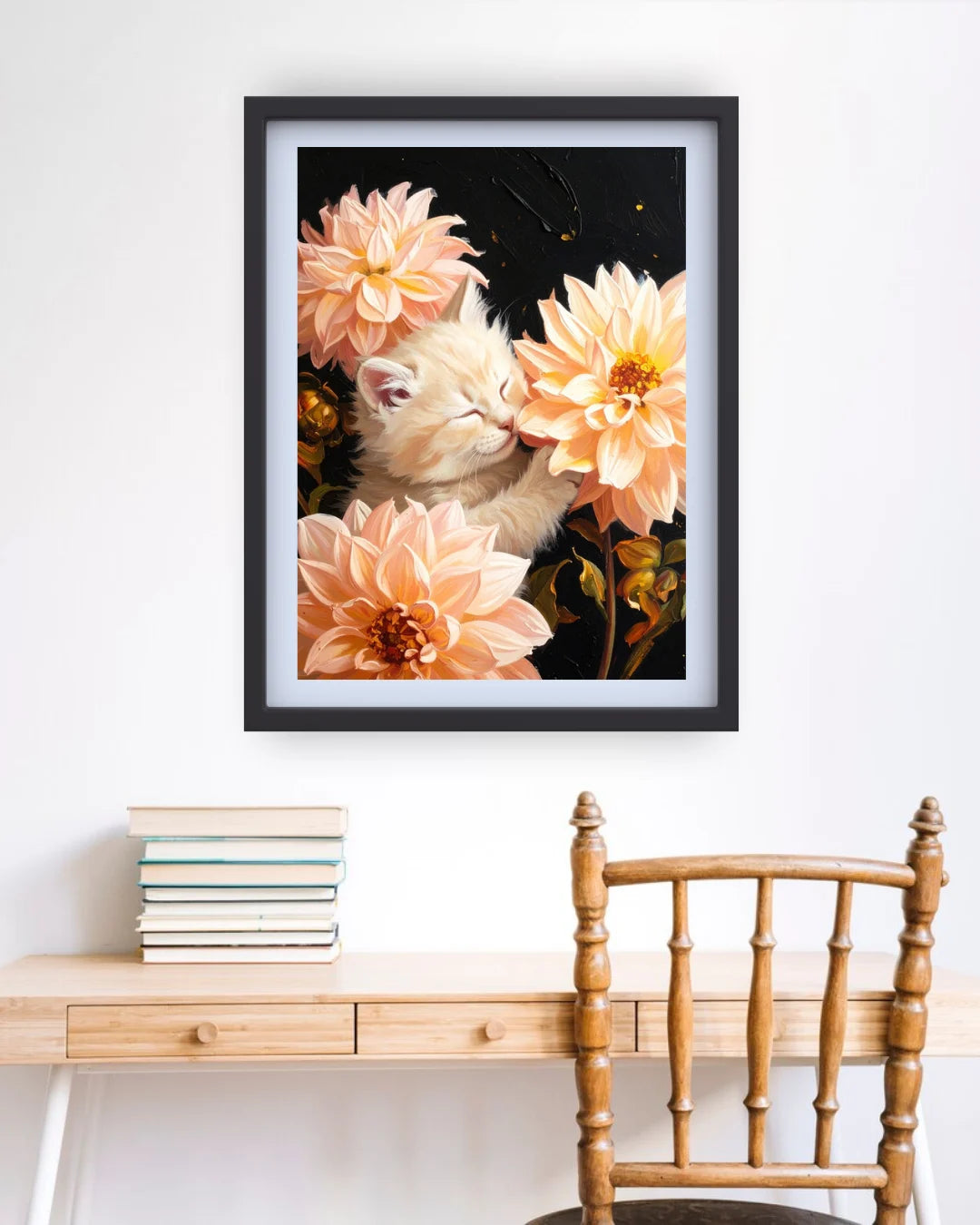 A framed art print featuring a fluffy cream-colored kitten nestled among peach and orange dahlia flowers against a dark background, hanging on a white wall above a wooden desk and chair.