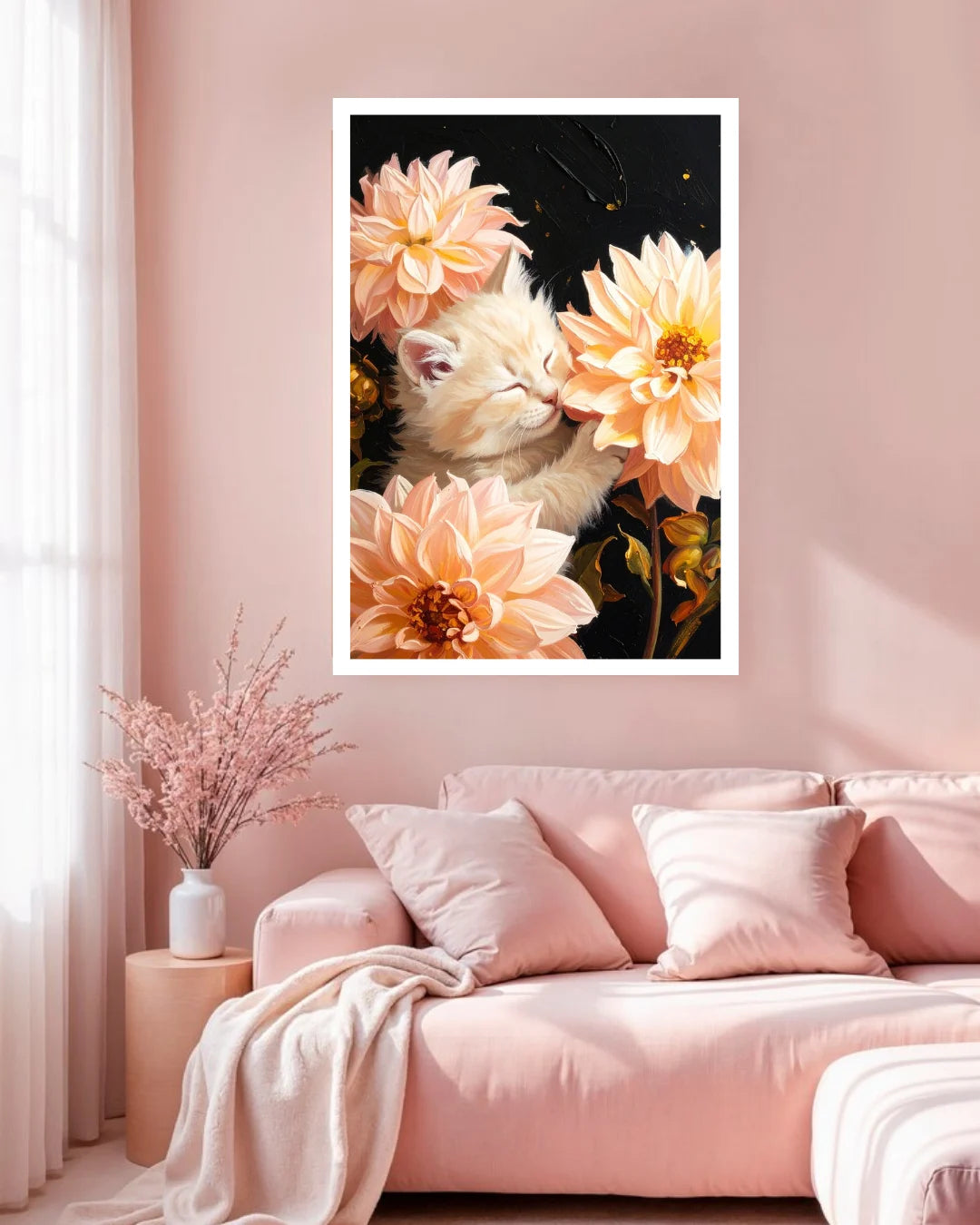 A framed wall art print featuring a cream-colored kitten nestled among large peach and pink dahlia flowers against a dark background, displayed above a pink sofa in a softly styled living room.