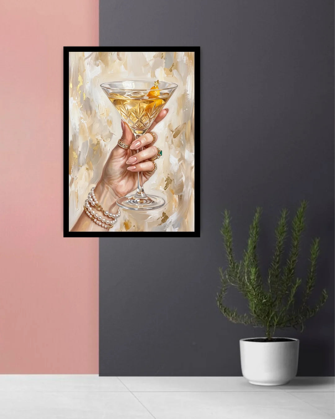 A framed art print depicting a hand holding a martini glass with an orange peel, displayed on a wall in a styled room setting with a pink and dark gray background and a potted plant.