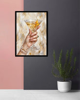 A framed art print depicting a hand holding a martini glass with an orange peel, displayed on a wall in a styled room setting with a pink and dark gray background and a potted plant.