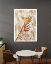 A framed art print depicting a hand holding a martini glass with a gold peel, displayed on a dark gray wall above a small round table and two chairs.