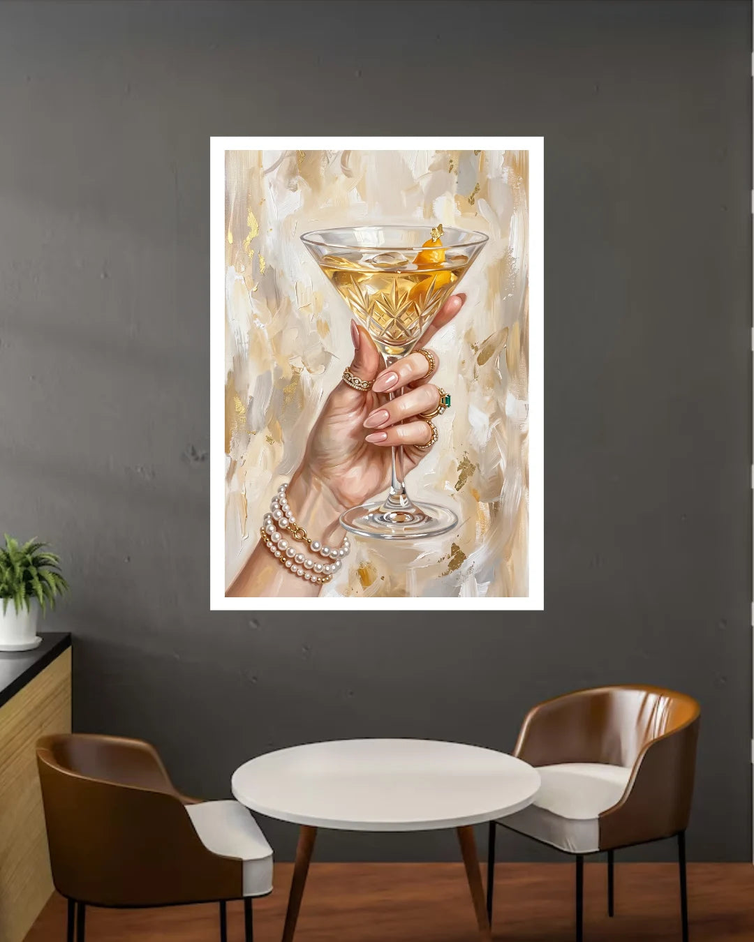 A framed art print depicting a hand holding a martini glass with a gold peel, displayed on a dark gray wall above a small round table and two chairs.