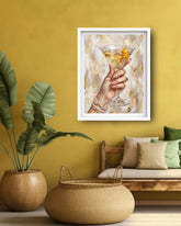 A framed art print depicting a hand holding a martini glass with a gold citrus twist, displayed on a mustard yellow wall in a styled living room setting with a bench, pillows, and a potted plant.