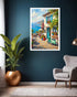 A framed vertical canvas print of a Mediterranean coastal cafe scene hangs on a dark blue wall in a modern living room setting with a gray armchair, a potted plant, and a floor lamp.