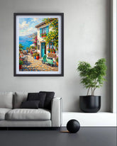 A framed art print depicting a colorful Mediterranean coastal village scene with a green scooter, blue sea, and white buildings, displayed on a light gray wall above a modern white sofa in a styled living room.