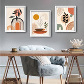 Three framed abstract botanical wall art prints in white frames hang above a wooden desk and a gray velvet armchair in a styled room setting.