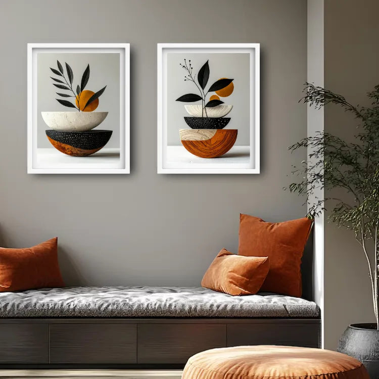 Two framed abstract botanical wall art prints hang above a cushioned bench in a styled interior. The prints feature stacked bowls and leaves in black, white, orange, and brown tones on a light gray background.