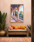 A framed art print depicting a colorful Mediterranean-style arched corridor with lush greenery, displayed on a warm beige wall above a woven rattan sofa with orange and pink pillows.