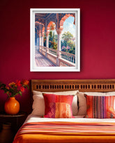 A framed art print depicting a vibrant Rajasthani-style archway corridor with colorful flowers and a blue sky, displayed on a deep red wall above a bed with orange and red patterned bedding.