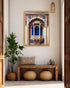 A framed art print depicting a traditional Indian Rajasthani chikankari jharoka arch with vibrant blue and gold details, displayed on a beige wall in a styled entryway with a wooden bench, potted plant, and woven decor.
