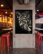 A framed black and white leopard art print hanging on a concrete pillar in a modern industrial bar with red stools and warm hanging lights.