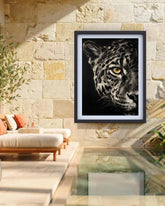 A framed black and white leopard art print with a yellow eye hanging on a beige stone wall above a modern outdoor lounge chair and glass pool.