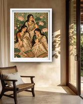 A framed art print depicting three women in traditional Indian sarees seated among lotus flowers, hung on a beige wall in a modern interior setting with a wooden chair and glass doors.