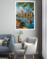 A framed wall art print depicting a scenic city harbor with a grand monument, boats on blue water, and green tree branches overhead, displayed above a gray armchair in a modern living room setting.