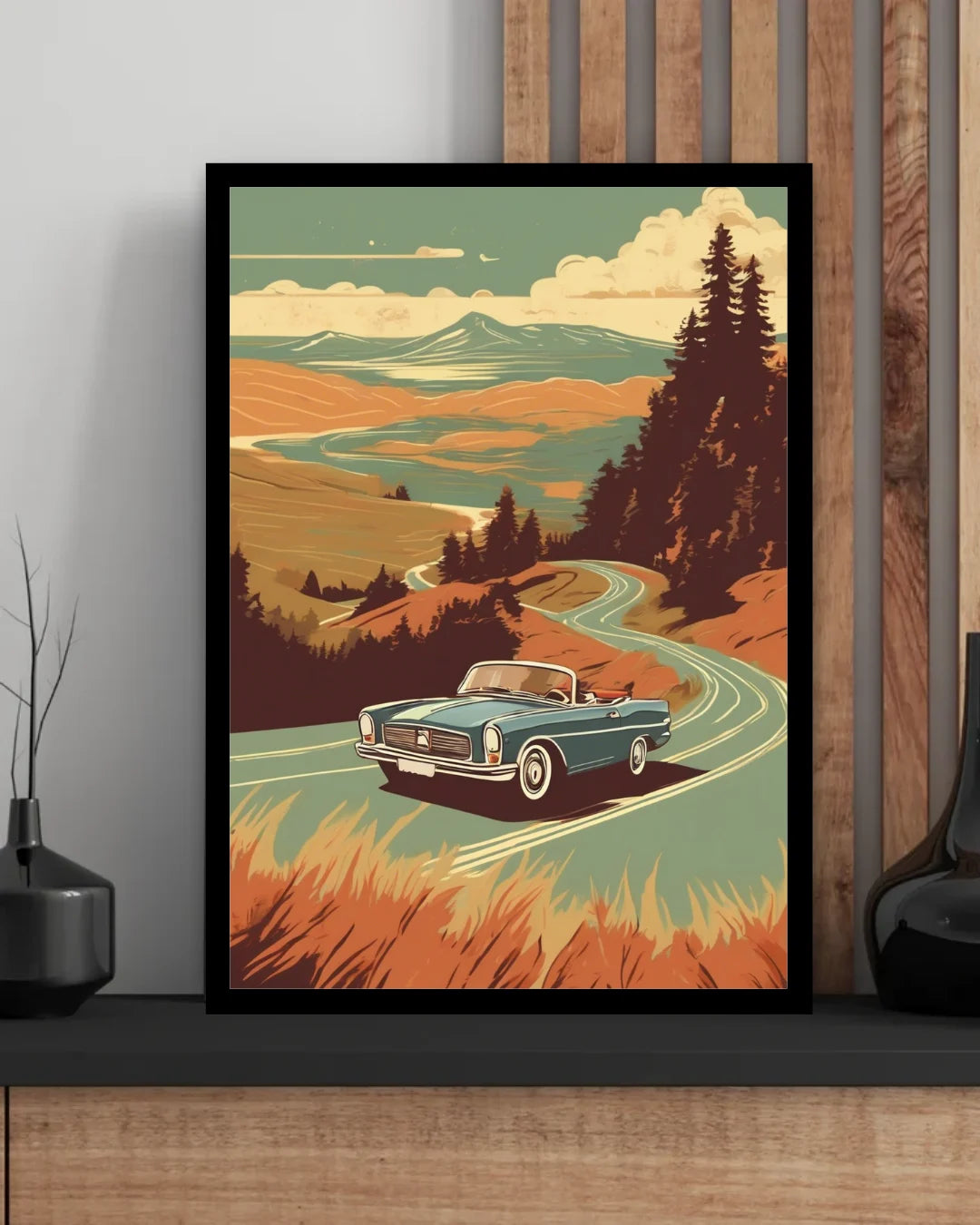 A framed art print featuring a retro illustration of a blue convertible car driving along a winding road through a stylized landscape, displayed on a shelf against a wood-paneled wall.